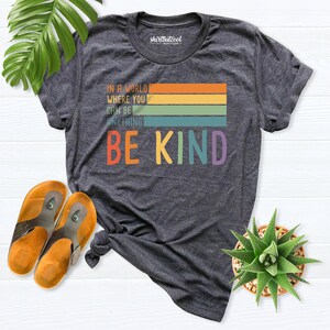 May include: A dark gray t-shirt with a rainbow stripe design and the text "In a world where you can be anything be kind".