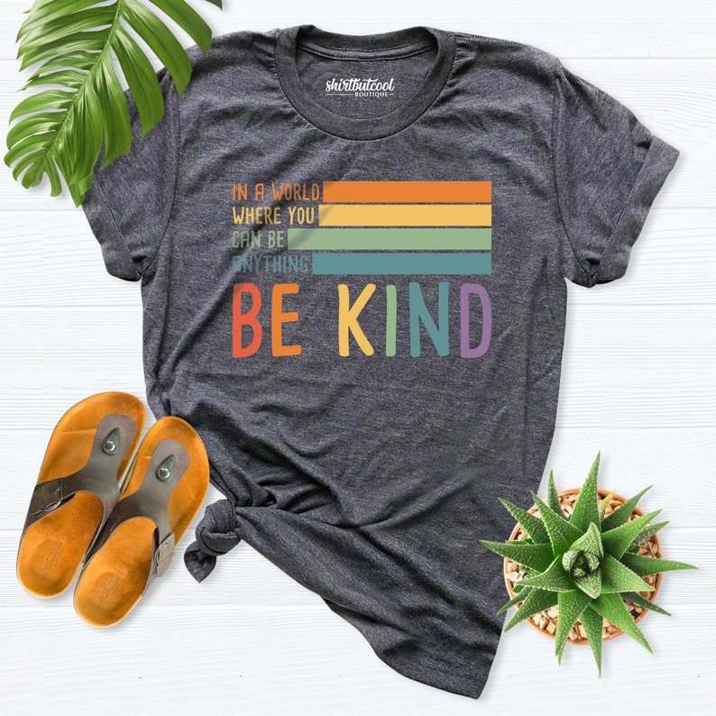 May include: A dark gray t-shirt with a rainbow stripe design and the text "In a world where you can be anything be kind".