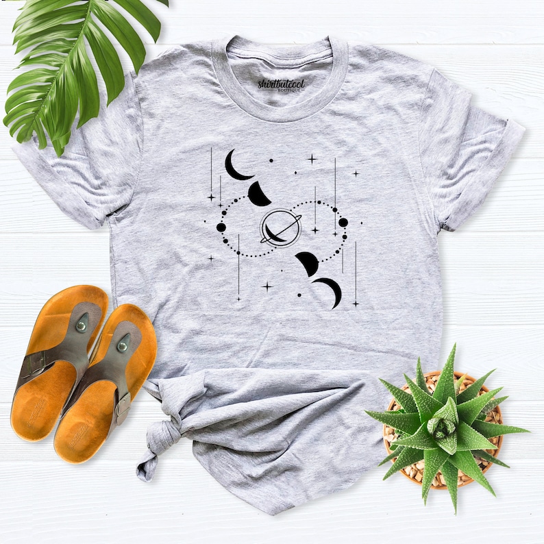 May include: A gray t-shirt with a black graphic design of a celestial scene with moons, stars, and a planet. The text "shirtatcool" is printed on the shirt.