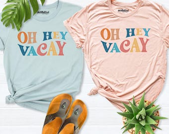 May include: A light blue t-shirt and a light pink t-shirt, both featuring the text OH HEY VACAY in multi-colored letters. The shirts are displayed alongside a pair of brown sandals and a green succulent plant.