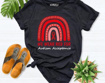 May include: A black short-sleeve t-shirt featuring a red rainbow graphic with a central red ribbon. The text reads WE WEAR RED FOR Autism Acceptance. The shirt has a crew neckline and is made of a solid black fabric with a white printed design.