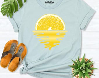 May include: A light blue t-shirt featuring a graphic of a yellow lemon slice positioned above a yellow reflection, with the text "shirtbutcool boutique" printed in black on the inside collar area of the garment.