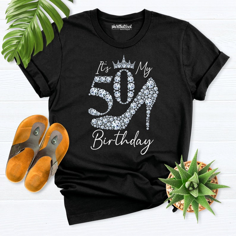 May include: Black t-shirt with the text "It's My 50th Birthday". The number 50 is designed with sparkling gems, and a high heel shoe is also made of gems. A crown is above the number 50.