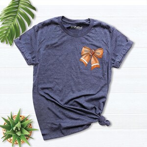 Pode incluir: Uma camiseta azul marinho com um la&ccedil;o em forma de bola de futebol marrom e laranja na frente. O la&ccedil;o tem as palavras "Game Day" escritas nele.