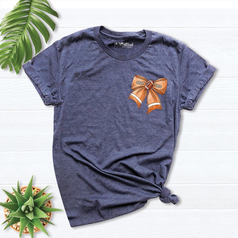 Pode incluir: Uma camiseta azul marinho com um la&ccedil;o em forma de bola de futebol marrom e laranja na frente. O la&ccedil;o tem as palavras "Game Day" escritas nele.