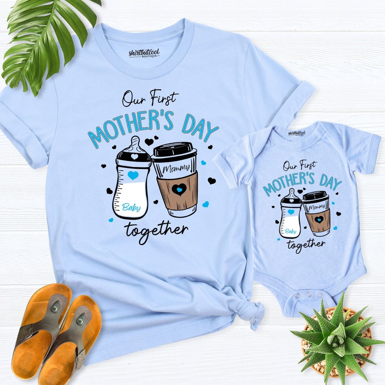 May include: A light blue t-shirt and onesie set with the text "Our First Mother's Day together" and illustrations of a baby bottle and a coffee cup. The baby bottle says "Baby" and the coffee cup says "Mommy".
