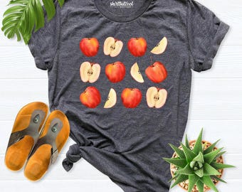 May include: A dark heather gray short-sleeve t-shirt featuring a graphic print of various whole and sliced red apples. The shirt has a crew neckline and the text shirtbutcool boutique printed in white on the inside collar area.