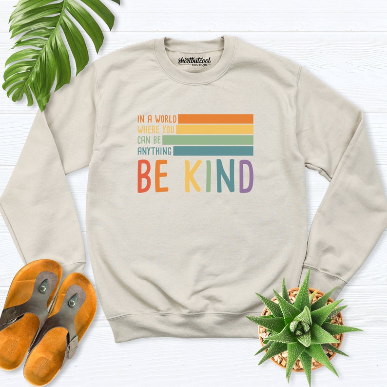 May include: A beige sweatshirt with the text "In a world where you can be anything be kind" in rainbow colors. The text is in a retro style font.
