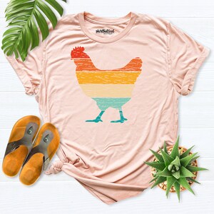 May include: A light pink t-shirt with a vintage-style graphic of a chicken. The graphic is in shades of orange, yellow, blue, and green. The shirt has a crew neck and short sleeves. The text "shirtbutcool" is printed on the tag.