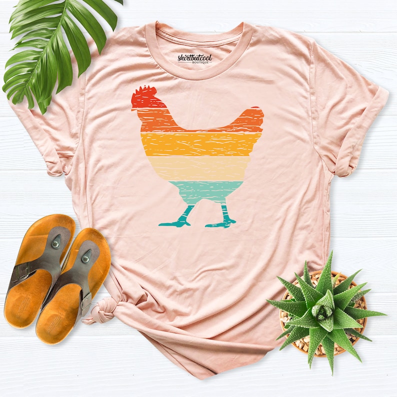 May include: A light pink t-shirt with a vintage-style graphic of a chicken. The graphic is in shades of orange, yellow, blue, and green. The shirt has a crew neck and short sleeves. The text "shirtbutcool" is printed on the tag.