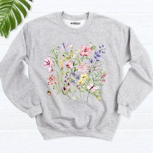 K&ouml;nnte beinhalten: Ein grauer Pullover mit einem Blumendruck mit rosa, gelben und blauen Blumen, gr&uuml;nen Bl&auml;ttern, einem Schmetterling und einer Libelle. Der Text "shirtbutcool" ist oben auf dem Pullover gedruckt.