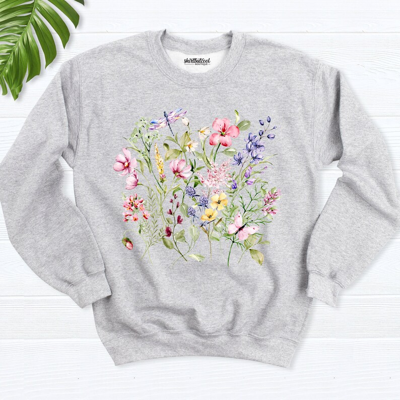 K&ouml;nnte beinhalten: Ein grauer Pullover mit einem Blumendruck mit rosa, gelben und blauen Blumen, gr&uuml;nen Bl&auml;ttern, einem Schmetterling und einer Libelle. Der Text "shirtbutcool" ist oben auf dem Pullover gedruckt.