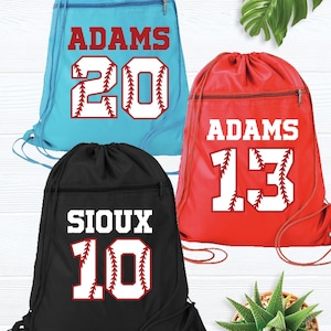 May include: Three drawstring sports bags in teal, red, and black. Each bag features a baseball-themed design with a name and number. The teal bag says "ADAMS 20", the red bag says "ADAMS 13", and the black bag says "SIOUX 10".