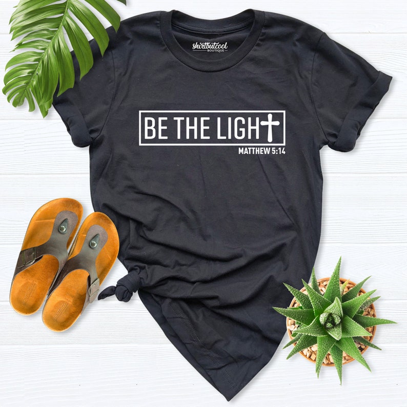 May include: Black t-shirt with white text that reads "BE THE LIGHT" with a cross symbol and "MATTHEW 5:14" underneath.