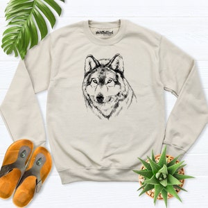 Pu&ograve; includere: Una felpa beige con un'illustrazione in bianco e nero di una testa di lupo sul davanti. Il testo "shirtbutcool" &egrave; stampato sulla parte superiore della felpa.