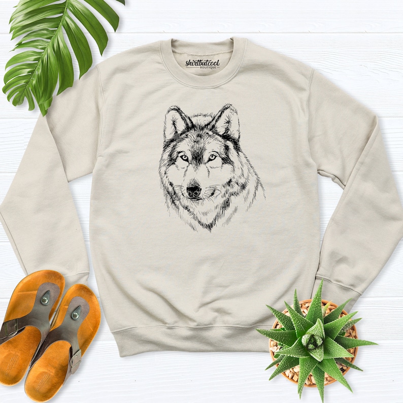 Pu&ograve; includere: Una felpa beige con un'illustrazione in bianco e nero di una testa di lupo sul davanti. Il testo "shirtbutcool" &egrave; stampato sulla parte superiore della felpa.