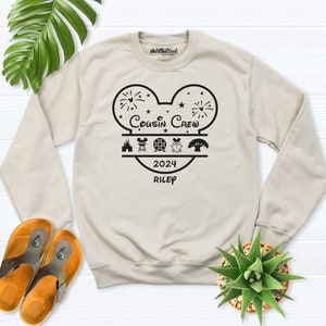 May include: A beige crewneck sweatshirt with a black graphic design. The design features a Mickey Mouse head shape with the words "Cousin Crew" and "2024 Riley". Below the text are icons of a castle, a building, a globe, and a tree. The sweatshirt is on a white wooden surface.