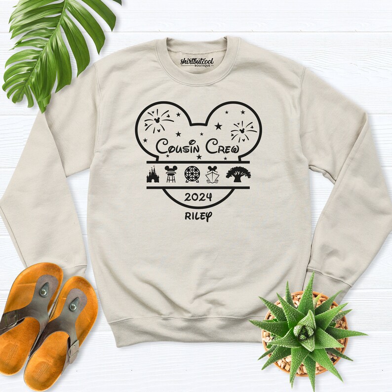 May include: A beige crewneck sweatshirt with a black graphic design. The design features a Mickey Mouse head shape with the words "Cousin Crew" and "2024 Riley". Below the text are icons of a castle, a building, a globe, and a tree. The sweatshirt is on a white wooden surface.