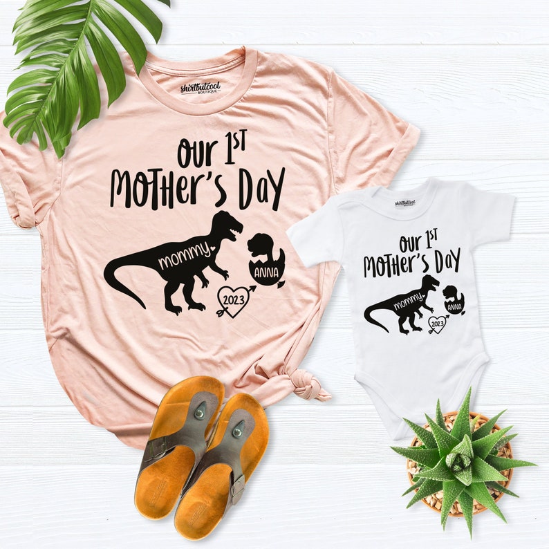 Peut inclure: Un t-shirt rose et un body blanc avec un motif de dinosaure et le texte "Our 1st Mother's Day". Le dinosaure sur le t-shirt dit "mommy" et le dinosaure sur le body dit "mommy". Le dinosaure sur le body a un plus petit dinosaure dans sa bouche qui dit "anna". Les deux t-shirts ont un c&oelig;ur avec l'ann&eacute;e 2023.
