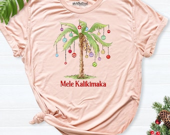 May include: A light peach-colored t-shirt featuring a graphic of a palm tree decorated with colorful hanging ornaments. Below the tree, the text "Mele Kalikimaka" is printed in red. The shirt has a crew neckline and short sleeves.