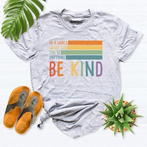 May include: A gray t-shirt with a rainbow stripe design and the text "In a world where you can be anything be kind".