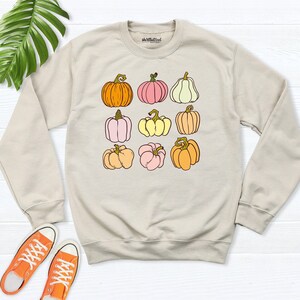 May include: A beige crewneck sweatshirt featuring a graphic print of nine pumpkins in various shades of orange, pink, yellow, and cream. The brand name "skwllbutcool" is printed in small black text on the inner neckline of the garment.
