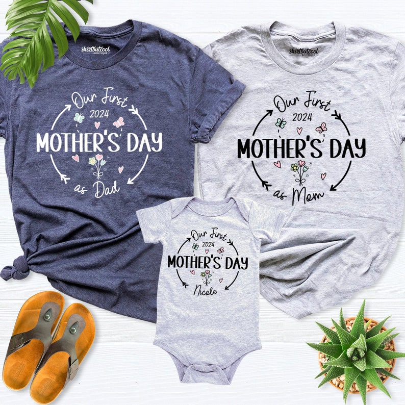 May include: Three grey t-shirts with a white graphic design that says "Our First Mother's Day" with a flower and heart design. The shirts are for a family of three, with the largest shirt saying "as Dad", the medium shirt saying "as Mum", and the smallest shirt saying "Nicole".