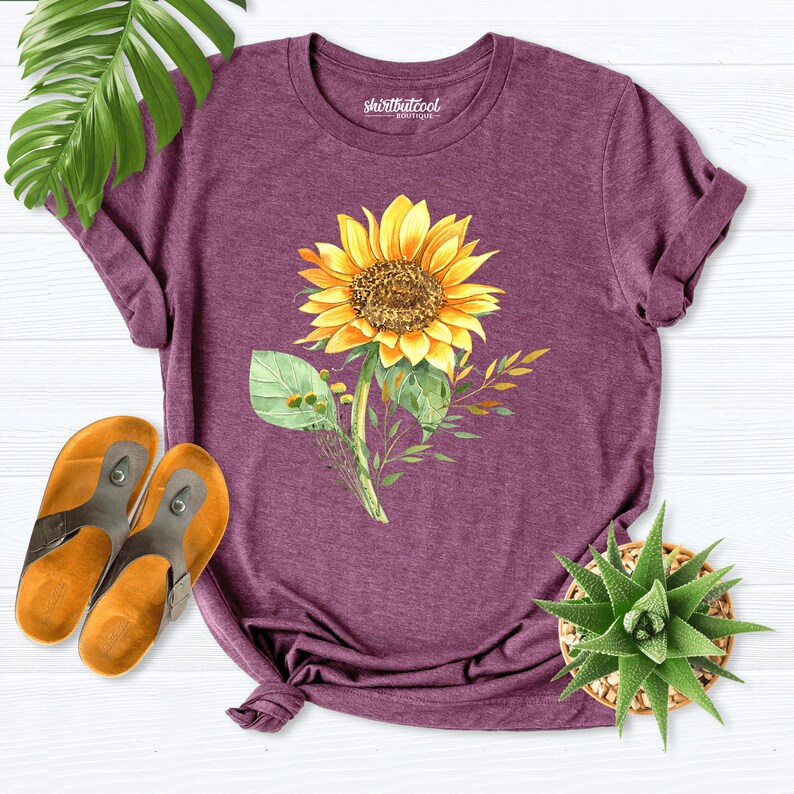 May include: A purple t-shirt with a watercolor sunflower design. The shirt has the text "shirtbutcool boutique" printed on the front.