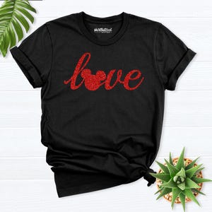 Pode incluir: Uma t-shirt preta de manga curta com a palavra love em letra cursiva de purpurina vermelha no peito, onde a letra o é substituída por uma silhueta de orelhas de rato. A etiqueta interior do pescoço apresenta o texto shirtbutcool boutique.
