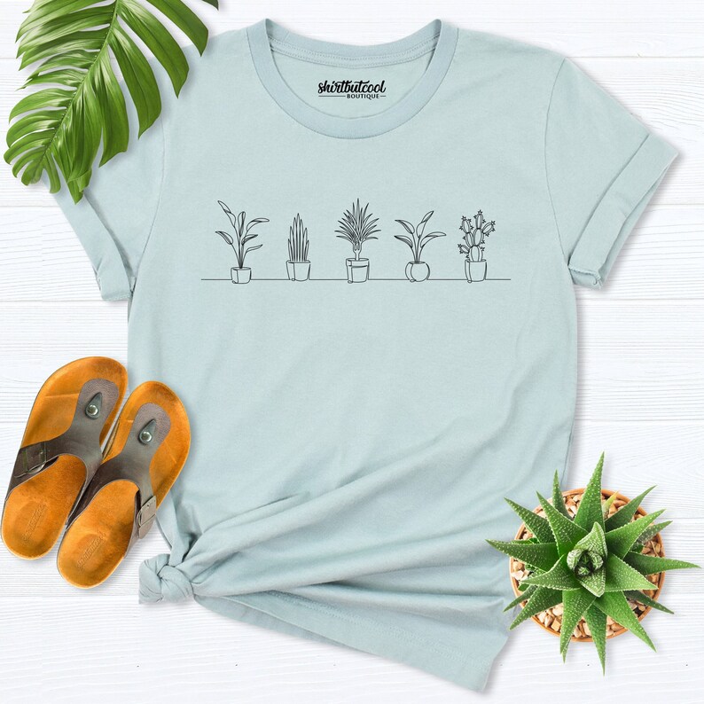 Puede incluir: Camiseta azul claro con un dibujo lineal de plantas en maceta. El texto "shirtbutcool boutique" est&aacute; impreso en la camiseta.