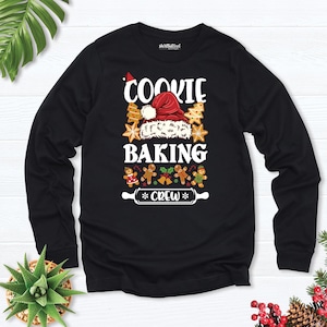 May include: A black long-sleeve shirt featuring a graphic print with the text "COOKIE BAKING CREW". The design includes a red Santa hat, various star-shaped cookies, gingerbread people, holly berries, and a rolling pin illustration.