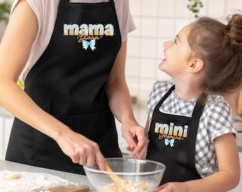 Puede incluir: Dos delantales negros con texto blanco y detalles en azul y naranja. El delantal más grande muestra la palabra mama y el nombre Diana con un lazo azul. El delantal más pequeño muestra la palabra mini y el nombre Jasmine con un lazo azul.