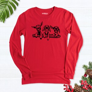 K&ouml;nnte beinhalten: Rotes Langarmshirt mit einer schwarzen Grafik von drei jungen Ziegen, die &uuml;ber einen Ast schauen.