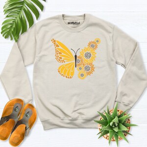May include: A beige sweatshirt with a yellow butterfly graphic made of sunflowers. The butterfly is centered on the sweatshirt and has a brown outline. The sweatshirt has the text "shirtbutcool" printed on the front.