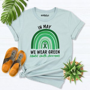 K&ouml;nnte beinhalten: Hellblaues T-Shirt mit einem gr&uuml;nen Regenbogen und einem gr&uuml;nen Band. Der Text "In May We Wear Green Mental Health Awareness" ist auf dem T-Shirt aufgedruckt.