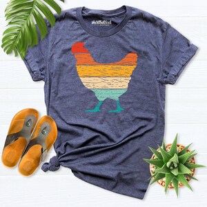 May include: A navy blue t-shirt with a vintage-style silhouette of a chicken in shades of orange, yellow, green, and blue. The shirt has the text "Shirtbutcool Boutique" printed on the front.