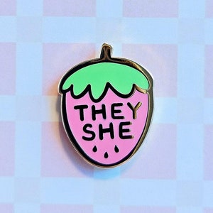 May include: A pink and green enamel pin shaped like a strawberry with the words "They She" inside. The pin is on a white and pink background with the text "misomomo" at the top and "misomomo.com" at the bottom.