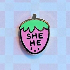 May include: A pink strawberry enamel pin with a gold outline. The strawberry has a green stem and leaves and the words "SHE HE" written on it. The pin is on a white and pink checkered background.