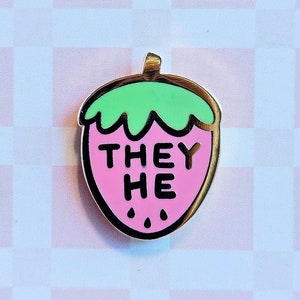 May include: Pink strawberry-shaped enamel pin with a gold outline. The strawberry has a green leafy top and the words "THEY HE" written on the front.