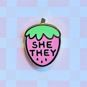 May include: A pink strawberry enamel pin with a gold outline. The strawberry has a green stem and leaves and the text "SHE THEY" in black letters.