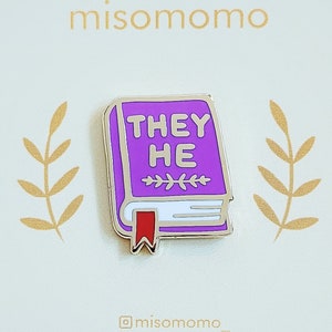 Peut inclure: Épinglette en émail violet en forme de livre avec les mots "They He" sur la couverture. L'épingle a un contour doré et un marque-page rouge.