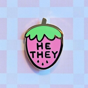 May include: A pink and green enamel pin shaped like a strawberry with the words "HE THEY" on it. The pin is on a white and pink checkered background.