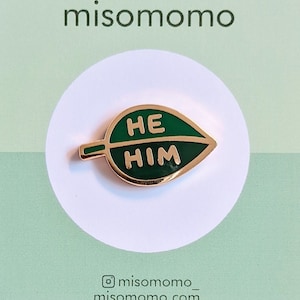 May include: A green leaf-shaped enamel pin with the words "HE HIM" in gold lettering. The pin is on a white card with the text "misomomo" and the social media handle "@misomomo_" and website "misomomo.com".