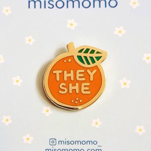 May include: Orange enamel pin with a gold outline and a green leaf. The orange has the words "They She" written on it. The pin is on a blue background with white flowers.