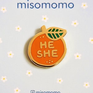 May include: Orange enamel pin with a gold outline and a green leaf. The orange has the words "HE SHE" written on it. The pin is on a light blue background with white flowers.