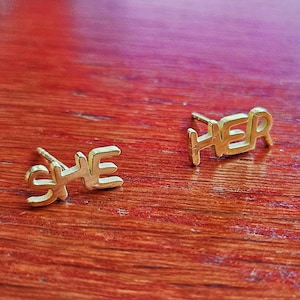 Könnte beinhalten: Ein Paar goldene Ohrringe mit den Worten "SHE" und "HER" darauf.