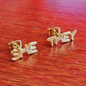 May include: Gold-colored stud earrings spelling out the word "SHE" and "THEY". The earrings are displayed on a reddish-brown wooden surface. The earrings are small and delicate, with a simple, modern design.