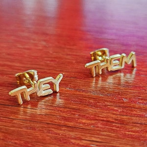 May include: A pair of gold-colored earrings with the words "THEY" and "THEM" spelled out in a stylized font.