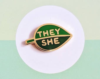 May include: Green enamel pin shaped like a leaf with the words "THEY SHE" in gold lettering.