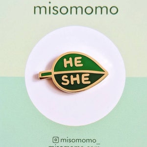 May include: A green leaf-shaped enamel pin with the words "HE" and "SHE" in white. The pin has a gold-colored border and is on a light green card with the brand name "misomomo". The card has a white circle behind the pin.